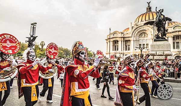 Desfile de músicos vestidos como Catrinas, comemorando o Dia dos Mortos no México, com o Palais de Bellas Artes ao fundo. Uma cena vibrante e cultural.