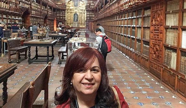 Deusa Rodrigues na Viagem para Puebla, em visita a uma biblioteca antiga com estantes de madeira recheadas de livros, arquitetura clássica e detalhes históricos ao fundo.