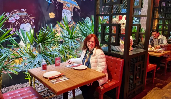Uma mulher está sentada à mesa de um restaurante, usando uma jaqueta clara e sorrindo para a câmera. O ambiente tem decoração colorida, plantas, painéis artísticos nas paredes e divisórias vazadas. Há pratos, talheres e outros itens sobre a mesa, enquanto outras pessoas estão sentadas ao fundo.