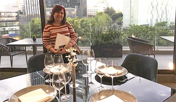 Deusa Rodrigues sorridente em um restaurante elegante com uma mesa bem arrumada, taças de vinho e com vista para a cidade ao fundo.