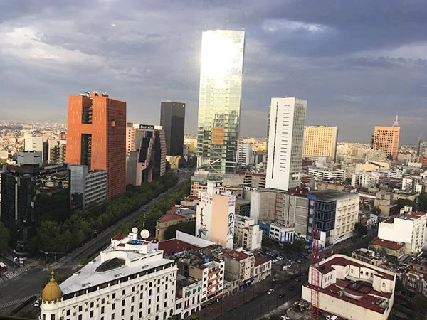 Imagem de arranha-céus modernos em um horizonte urbano de uma cidade grande, com destaque para um edifício de vidro refletivo ao centro e céu nublado ao fundo.