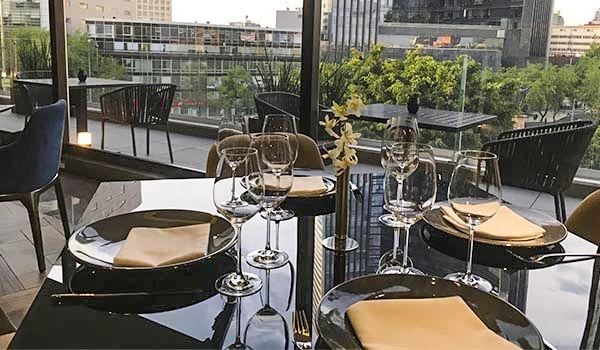 Imagem de uma mesa de jantar elegantemente preparada para um jantar sofisticado em um restaurante com vista para a cidade, incluindo taças de vinho, pratos e guardanapos.