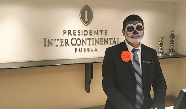 Homem vestido de terno com maquiagem de caveira mexicana em evento no Presidente Intercontinental Puebla, participando de celebração ou cerimônia.