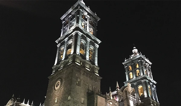 Imagem das torres da Catedral de Puebla iluminadas à noite, destacando detalhes arquitetônicos e o céu escuro ao fundo.