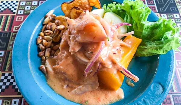 Prato de comida peruana servido em um prato azul. No centro, há peixe em pedaços coberto por um molho cremoso, acompanhado de cebola roxa, pimentão e outros vegetais. Ao redor, aparecem milho torrado, batata ou mandioca frita, legumes e uma folha de alface. O prato está sobre uma toalha de mesa com estampa geométrica colorida.