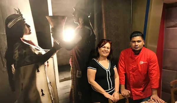 Fotografia de três pessoas em frente a um mural com temática indígena. Uma mulher e um homem estão lado a lado, sorrindo para a câmera, enquanto uma representação de uma figura indígena aparece no mural ao fundo.