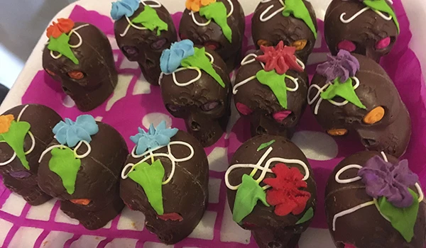 Imagem de doces em formato de caveira, decorados com glacê colorido em tons de azul, verde, rosa, roxo e laranja, com cobertura de chocolate escuro, típicos do Dia dos Mortos.