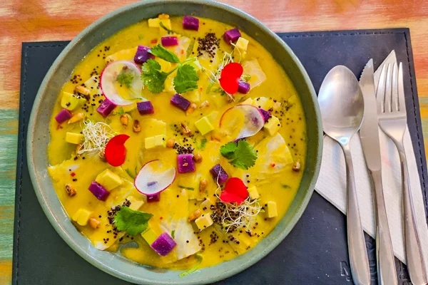 Prato peruano servido em um prato verde, com uma preparação de molho amarelo coberta por fatias finas de peixe e decorada com rodelas de rabanete, cubos de batata-roxa e milho, brotos, folhas de coentro, sementes e pequenas pétalas vermelhas. Ao lado, há uma colher e um garfo sobre um guardanapo branco.