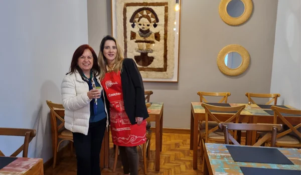 Duas mulheres posam sorrindo em um restaurante. À esquerda, uma mulher usa casaco branco e segura uma taça; à direita, outra veste roupa preta e um avental vermelho com referências à culinária peruana. Ao fundo, há mesas e cadeiras de madeira, uma obra de arte com figura humana e dois espelhos circulares na parede.