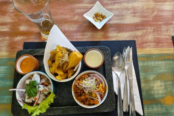 Mesa de restaurante peruanao centro SP, com um conjunto de pratos da culinária peruana servido sobre uma bandeja preta. Há um ceviche com peixe, cebola roxa, coentro e folhas de alface; um prato com pedaços fritos e acompanhamentos; e outro com uma preparação à base de legumes, cebola e brotos. Também há dois pequenos recipientes com molhos, talheres e um copo de bebida sobre a mesa de madeira.