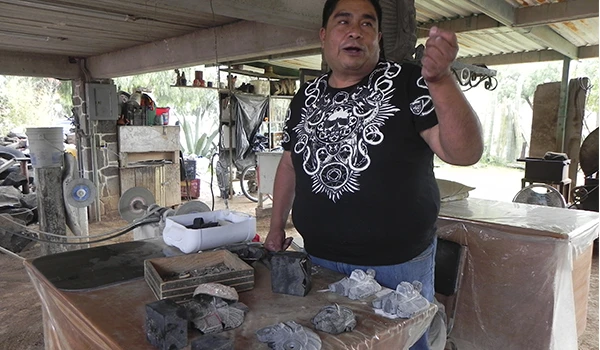 Homem explicando história ou cultura amazônica em um ambiente rústico, com objetos indígenas e entusiastas do artesanato local ao fundo.
