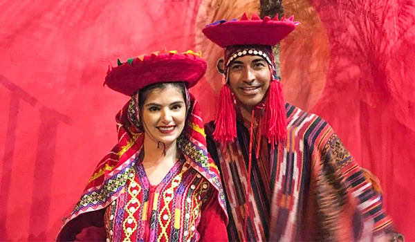 Duas pessoas posam sorrindo diante de um fundo vermelho, usando trajes tradicionais peruanos coloridos. A mulher, à esquerda, veste uma roupa vermelha com padrões geométricos e um chapéu ornamentado com franjas. O homem, à direita, usa uma túnica listrada em várias cores, chapéu decorado e longas faixas vermelhas ao redor do rosto. Restaurante peruano Centro SP.