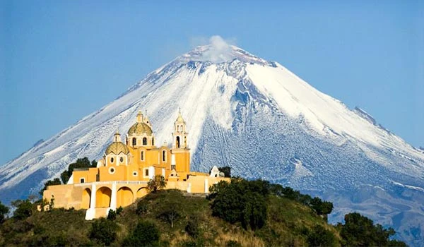 Imagem do vulcão como pano de fundo, com uma igreja mexicana colorida na frente, em um cenário natural exuberante, ideal para quem busca destinos turísticos no México.