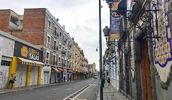 Viagem para Puebla, foto de uma rua com edifícios históricos, lojas e calçada de pedra, dia nublado, atmosfera urbana tranquila e cultural.