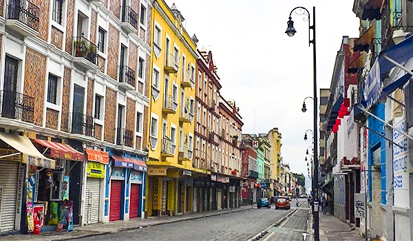 Imagem de uma rua no centro de uma cidade com edifícios coloridos e lojas fechadas, com carros estacionados ao longo da via, durante o dia.