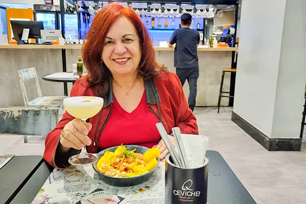 Deusa Rodrigues, sentada à mesa em um restaurante, segurando uma taça de bebida cremosa. À sua frente há um prato de ceviche com milho, cebola e pedaços de batata-doce, além de um recipiente com talheres. Ao fundo, vê-se o balcão e a área de atendimento do restaurante.