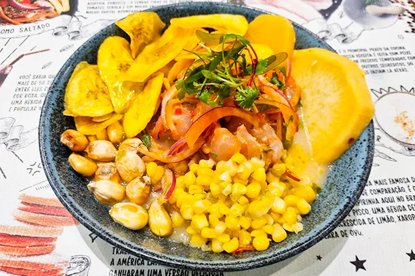 Prato de ceviche servido em prato escuro, acompanhado de banana-da-terra frita, batata amarela cozida, milho, milho torrado e cebola roxa. O ceviche é decorado com ervas frescas e tiras de cebola e pimentão, sobre uma mesa com papel estampado.
