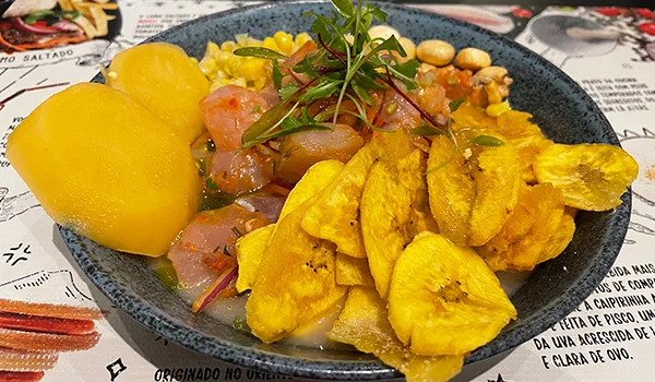 Prato de ceviche servido em um prato escuro, acompanhado de rodelas de banana-da-terra frita, batatas amarelas cozidas, milho e cebola roxa. O ceviche é decorado com ervas frescas, e o prato está sobre uma mesa com papel impresso.