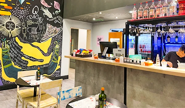 Interior de um restaurante com decoração contemporânea. À esquerda, há um grande mural colorido com ilustrações e referências à culinária peruana, incluindo a palavra “CEVICHE”. Ao centro e à direita, um balcão de atendimento com caixa, utensílios e bebidas expostas em uma estante refrigerada. Uma pessoa trabalha atrás do balcão. Na parte inferior, há mesas e cadeiras para os clientes.
