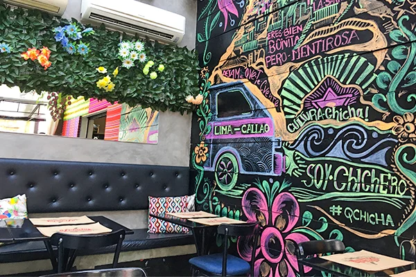 Restaurante com parede decorada com grafites coloridos, desenhos artísticos e frases em letras estilizadas. O ambiente possui bancos estofados pretos, mesas pequenas, almofadas estampadas e uma parede verde com folhagens e flores artificiais, criando um espaço descontraído e criativo.