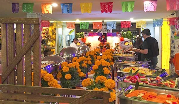 Decoração de festa com flores de cempasuchil e decoração colorida em ambiente festivo com pessoas servindo comida.
