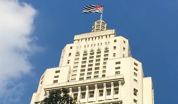 Prédio alto e branco, de arquitetura modernista, visto de baixo contra um céu azul. No topo, há uma torre com uma bandeira listrada em preto e branco e detalhes em vermelho.