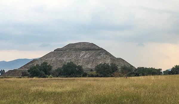 Imagem de uma das Pirâmides do Sol e da Lua no México, rodeada por vegetação e céu nublado, ideal para turismo e história.