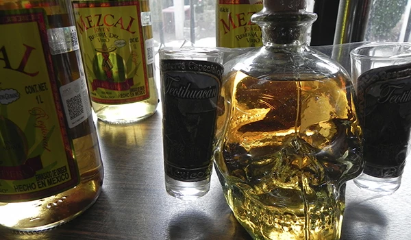 Imagem de garrafas de tequila e copos transparentes na mesa, com destaque para uma garrafa com formato de caveira de tequila, ideal para apreciadores de bebidas mexicanas.