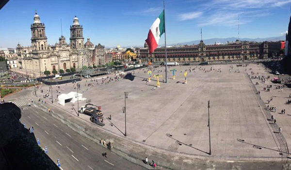 Praça principal na cidade do México com a bandeira mexicana ao centro, ao fundo a arquitetura colonial dos edifícios históricos e céu azul claro.