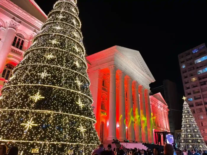 Árvore de Natal decorada com luzes brilhantes em uma praça urbana, ao lado de um prédio clássico iluminado, durante a noite.