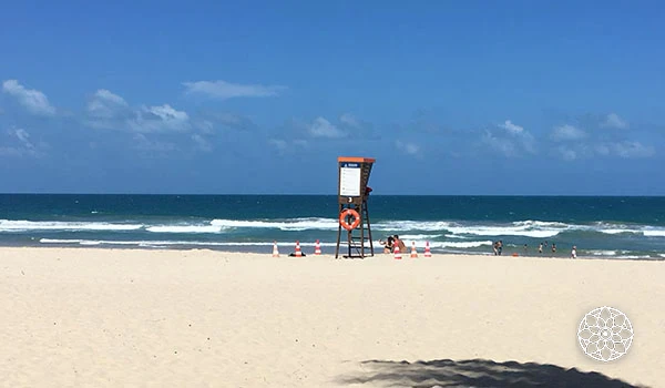 Praia de areia dourada com mar azul claro, céu com nuvens brancas, cabide de salva-vidas e algumas pessoas curtindo o dia de sol na orla.