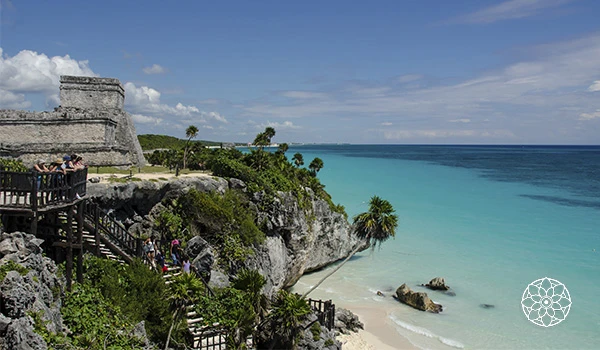 Paisagem de praia no México com falésias, mar azul claro, vegetação tropical e ruínas antigas ao fundo, representando o litoral turístico de Cancun.