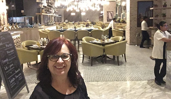 Deusa Rodrigues em restaurante elegante, com ambiente bem decorado, mesas bem arrumadas e funcionários preparando o espaço, ideal para experiências gastronômicas.