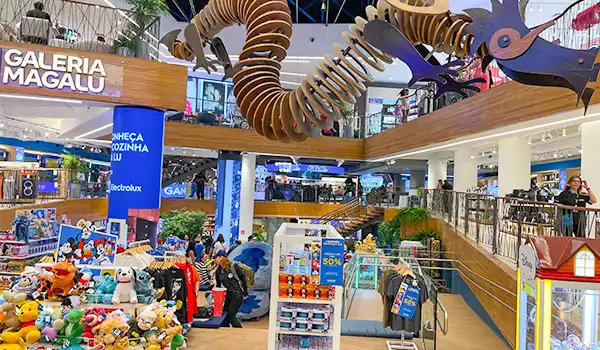 Visão de um shopping center com brinquedos, lojas e uma decoração infantil com um grande escorregador de tubo na parte superior, destacando a movimentação de pessoas.