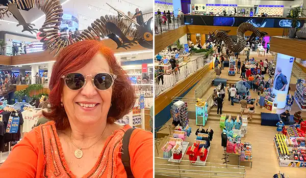 Mulher com cabelo castanho, óculos escuros e camiseta laranja posando no shopping com decoração de escorpião gigante no teto, e vista do interior de loja com várias pessoas e itens de venda.