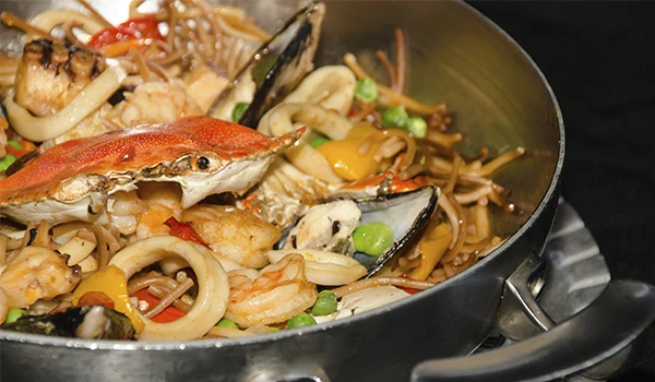 Imagem de uma panela com frutos do mar, incluindo caranguejo, camarões, lula e mexilhões, acompanhados de legumes como pimentão vermelho, ervilhas e cebolas, preparados em um prato de frutos do mar.