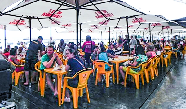 Imagem de um movimento de pessoas em uma feira ao ar livre sob grandes guarda-sóis brancos, com mesas e cadeiras de plástico amarelas, desfrutando de convívio social.