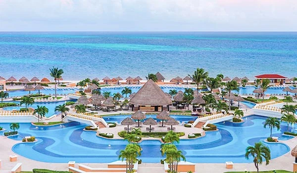 Vista panorâmica de resort de praia com piscinas, palmeiras e cabanas à beira-mar na costa de um destino turístico tropical, com mar azul ao fundo.