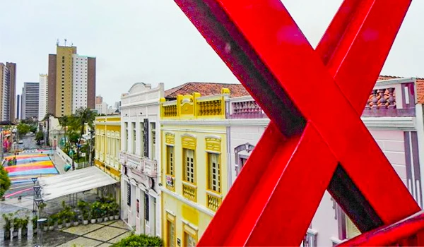 Imagem de uma rua vibrante de uma cidade com prédios coloridos, incluindo construções amarelas e brancas, ao lado de uma instalação artística vermelha com linhas diagonais. Fortaleza o que fazer.