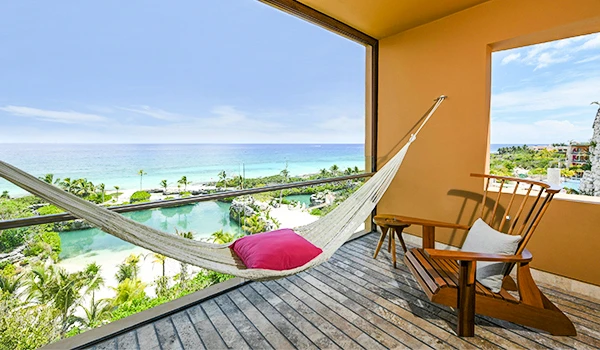Varanda com vista para mar cristalino e área de lazer com rede, cadeiras de madeira, clima ensolarado e paisagem tropical ao fundo.
