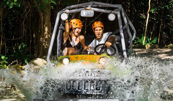 Aventura de lazer com duas pessoas em um veículo off-road atravessando um rio em meio à natureza, com capacetes de segurança e expressões de emoção e diversão. Para jovens casais na Lua de Mel em Cancun.