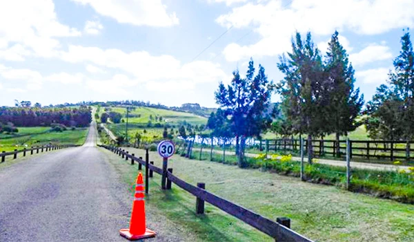 Imagem de uma estrada rural com sinal de limite de velocidade de 30 km/h, cones de trânsito, cercas de madeira e árvores verdes sob céu parcialmente nublado.