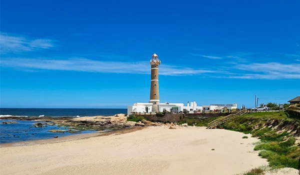 Farol na costa de uma praia ensolarada, com céu azul claro e mar calmo, ideal para turismo e viagens costeiras.