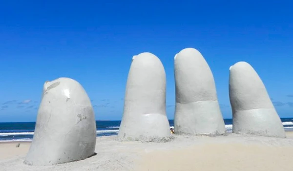 Esculturas de mãos gigantes feitas de areia na praia, com fundo de céu azul claro e mar ao fundo, formando uma instalação artística única na areia. Cassino Punta del Este.
