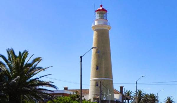 Foto de um farol em céu azul ao fundo e vegetação.
