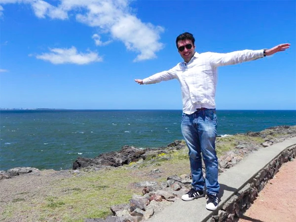 Homem sorridente com braços abertos na orla da praia, aproveitando o clima ensolarado e o mar ao fundo, em um dia claro e bonito.