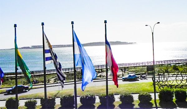 Imagem de várias bandeiras ao vento na orla de uma cidade com mar ao fundo, céu claro e trânsito com carros na rua. Um cenário urbano com aspecto de litoral. Cassino Punta del Este.