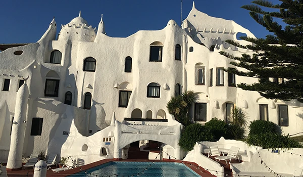 Fachada de uma construção arquitetônica moderna e única, com detalhes arredondados e cores brancas, ao lado de uma piscina e árvores verdes, ideal para férias na praia. Cassino Punta del Este.