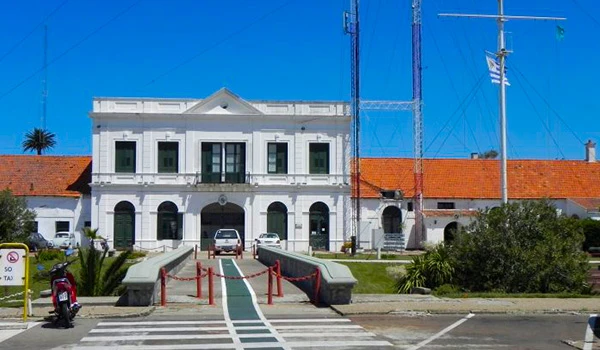 Imagem da fachada de um prédio branco com arquitetura clássica, com acesso por uma rampa e estacionamento na frente, sob céu azul claro.