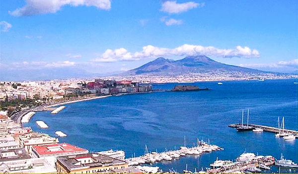 Vista panorâmica do litoral de Nápoles, com a baía, barcos e mar azul em primeiro plano e o Monte Vesúvio ao fundo.
